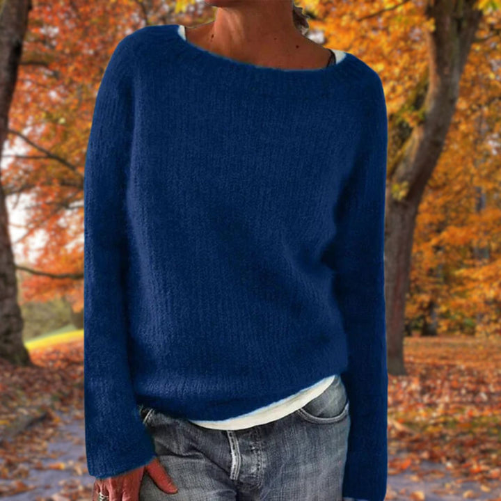 Kvinde i mørkeblå strikket sweater med rund hals, båret over hvid trøje og jeans i efterårsskov. 3