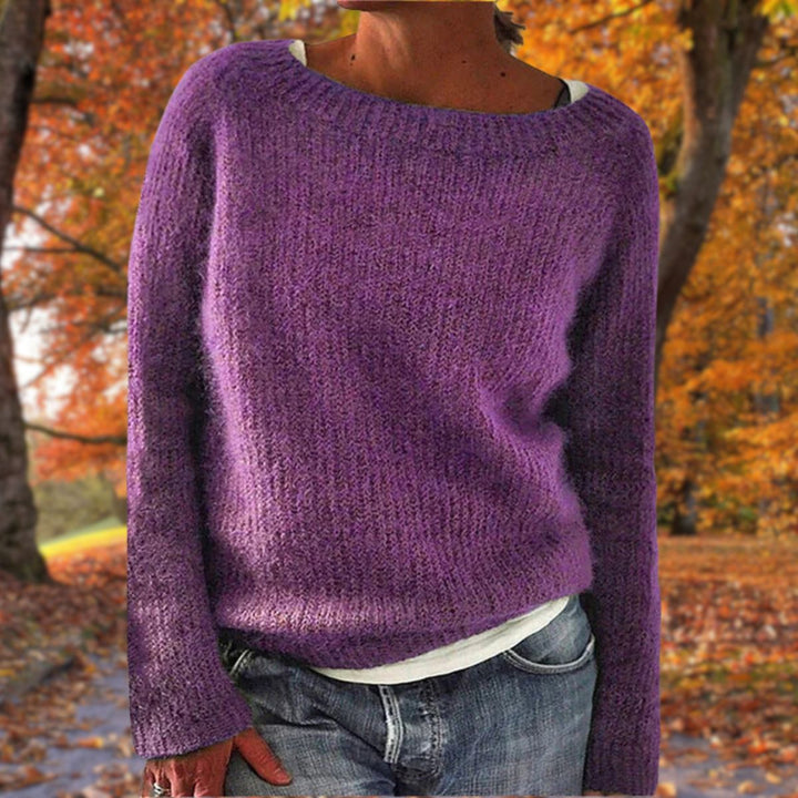 Kvinde i lilla strikket sweater med rund hals, båret over hvid trøje og jeans i efterårsskov. 7