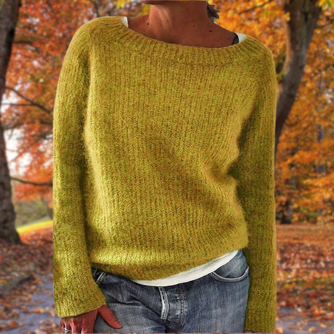 Kvinde i sennepsgul strikket sweater med rund hals, båret over hvid trøje og jeans foran efterårsløv. 1