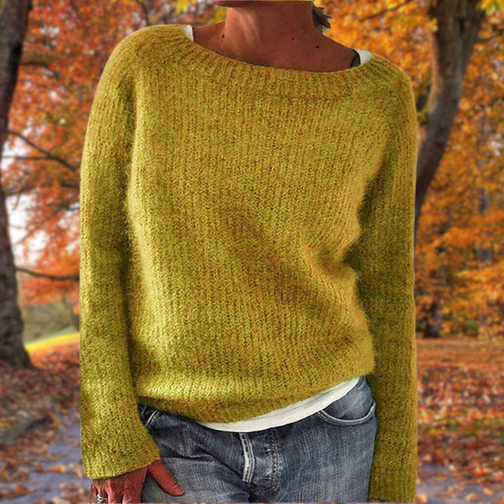 Kvinde i sennepsgul strikket sweater med rund hals, båret over hvid trøje og jeans foran efterårsløv. 1