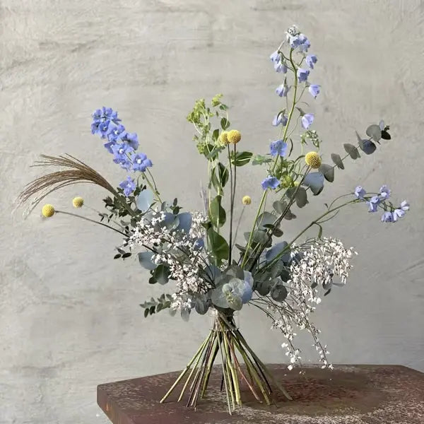 Frit stående buket med blå rittersporer, gule trommestikblomster, eukalyptus og hvide smådele foran en grå betonvæg. 17
