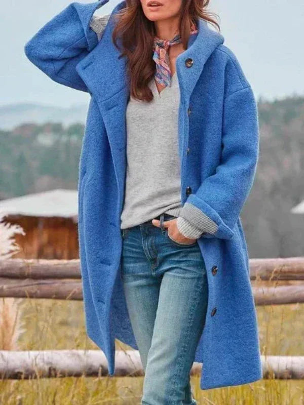 Kvinde i blå lang frakke med hætte og store knapper, båret åben over grå trøje og jeans. 4