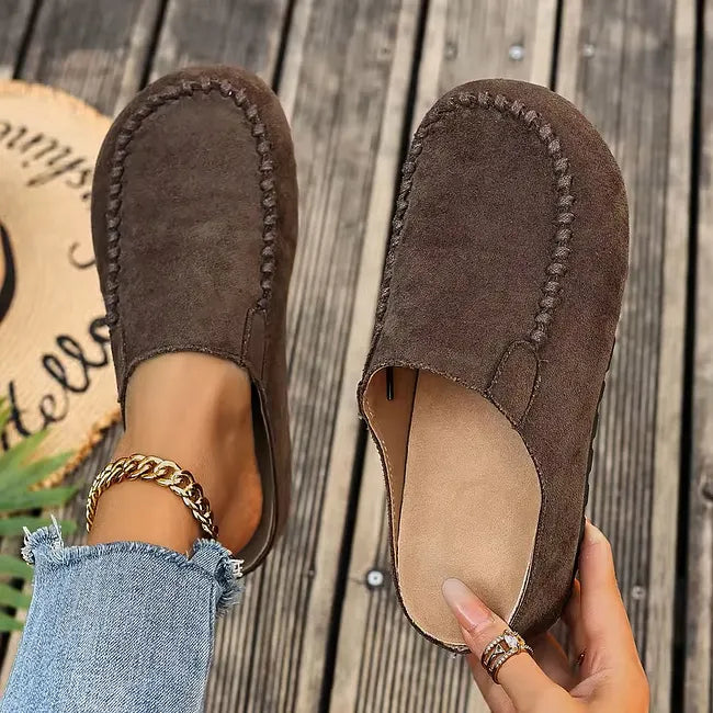 Mørkebrune ruskindsagtige slip-ons set ovenfra, den ene på foden og den anden løftet i hånden med lys indersål synlig. 3