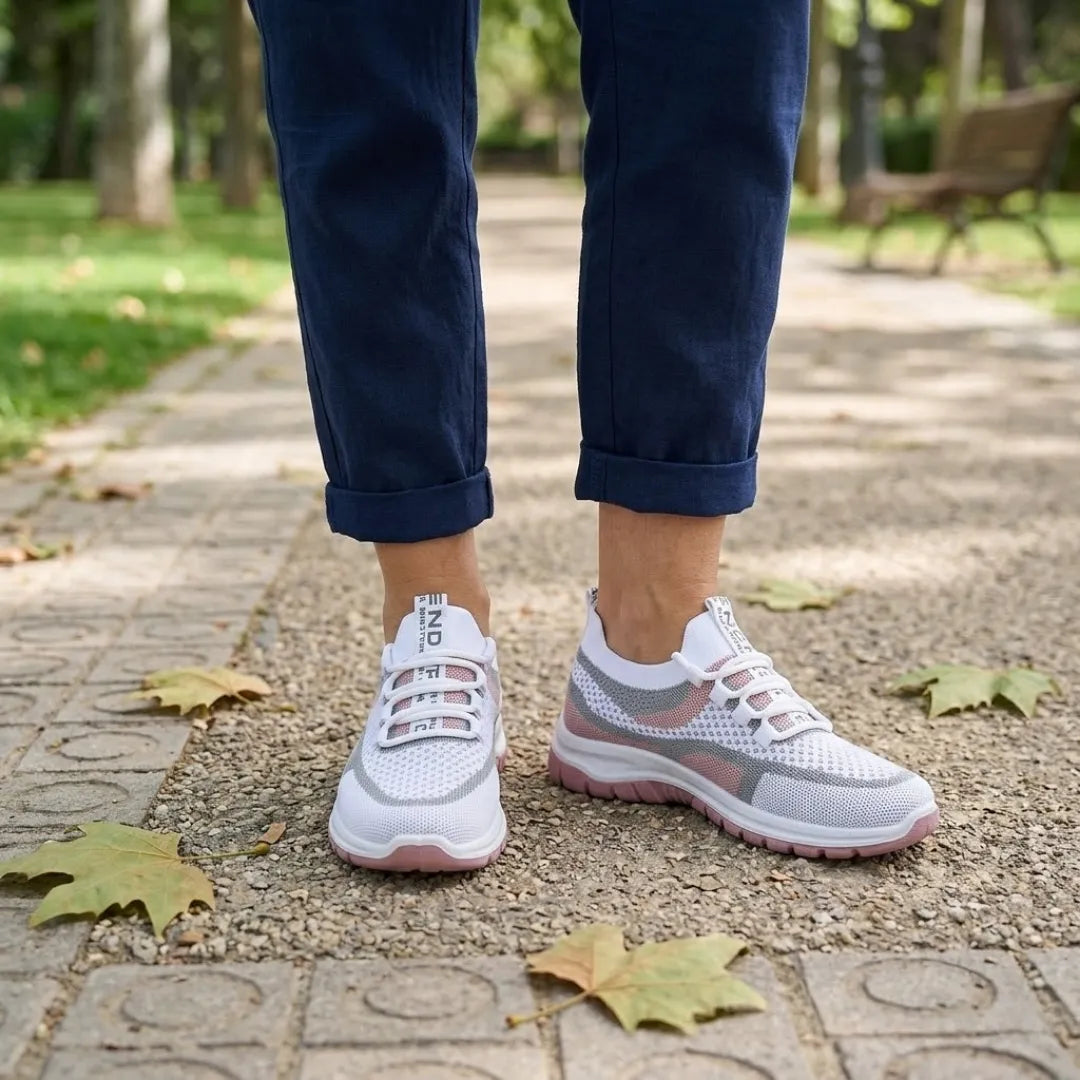Hvide sneakers med rosa sål båret af en kvinde i mørkeblå bukser på en sti i en park. 20
