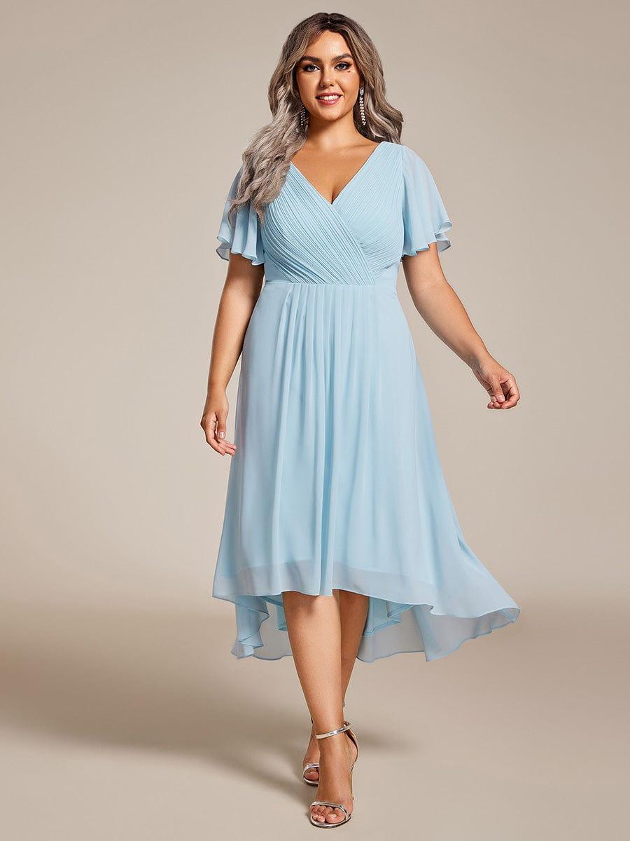 Plus size-model i lyseblå chiffonkjole med plisseret V-hals og asymmetrisk fald, set forfra med sølvfarvede sandaler. 89