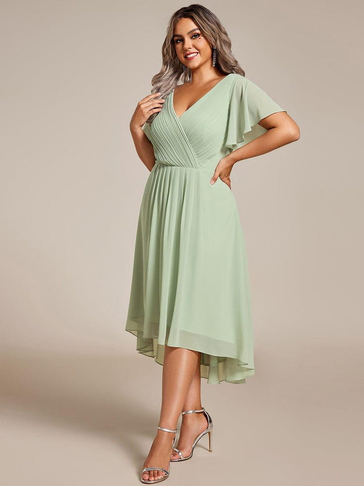 Model i plus size iført mintgrøn chiffonkjole med plisseret V-hals og asymmetrisk fald, set skråt forfra med hånd på hoften. 81