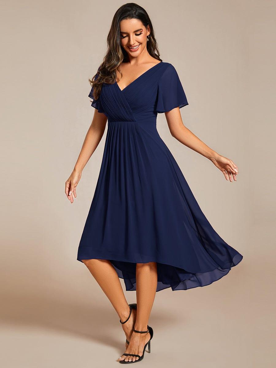 Model i marineblå chiffonkjole med V-hals ser smilende nedad, mens nederdelen svinger og den asymmetriske kant ses. 52