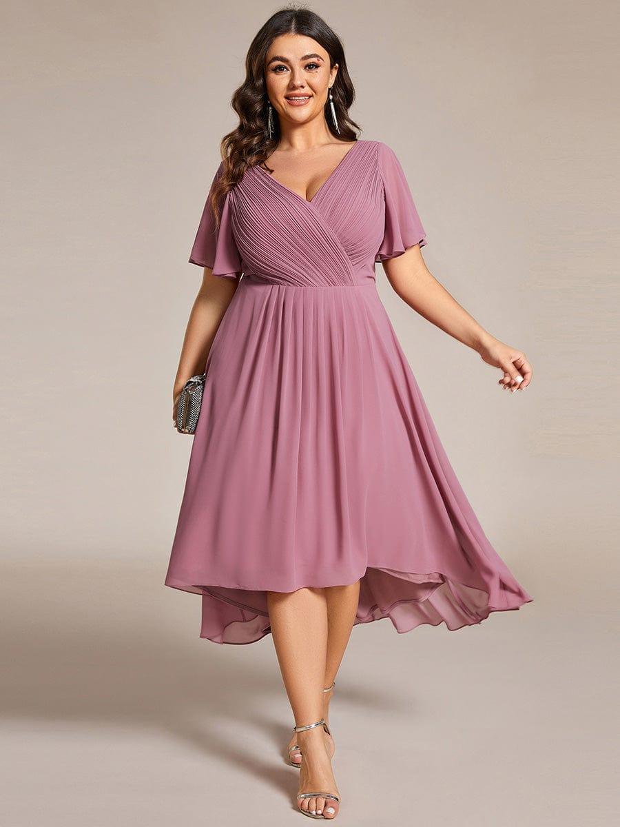 Plus size-model i mauve kjole med V-hals, korte flagrende ærmer og asymmetrisk fald, holder en lille clutch. 63