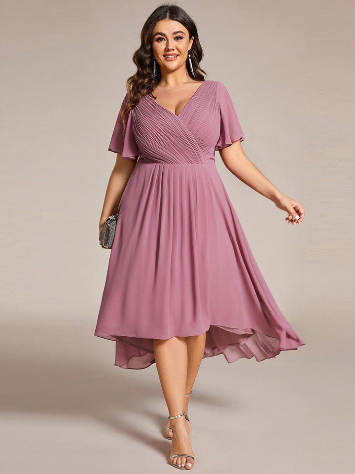 Plus size-model i mauve kjole med V-hals, korte flagrende ærmer og asymmetrisk fald, holder en lille clutch. 63