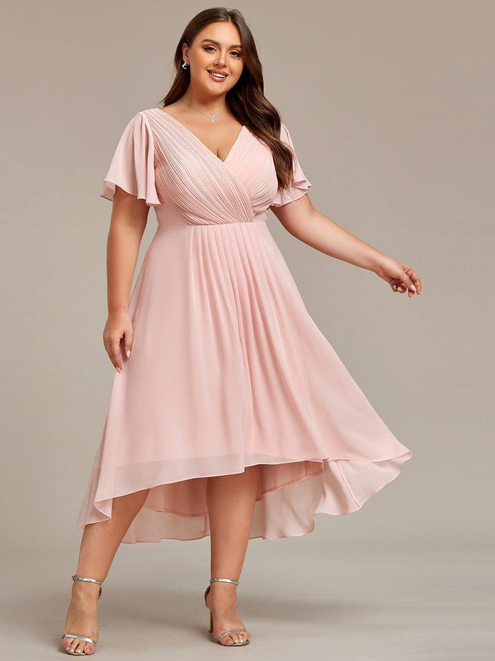 Lyserød kjole i plussize set forfra på smilende model med asymmetrisk fald og sølvfarvede sandaler. 34