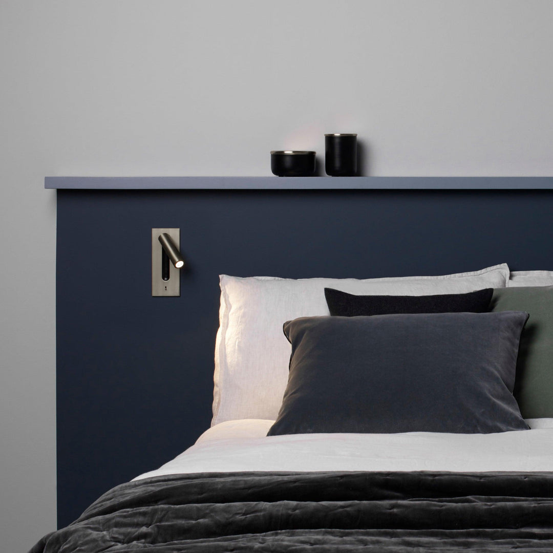 Væglampe i dæmpet metalfinish over en seng med blå sengegavl og grå puder 30