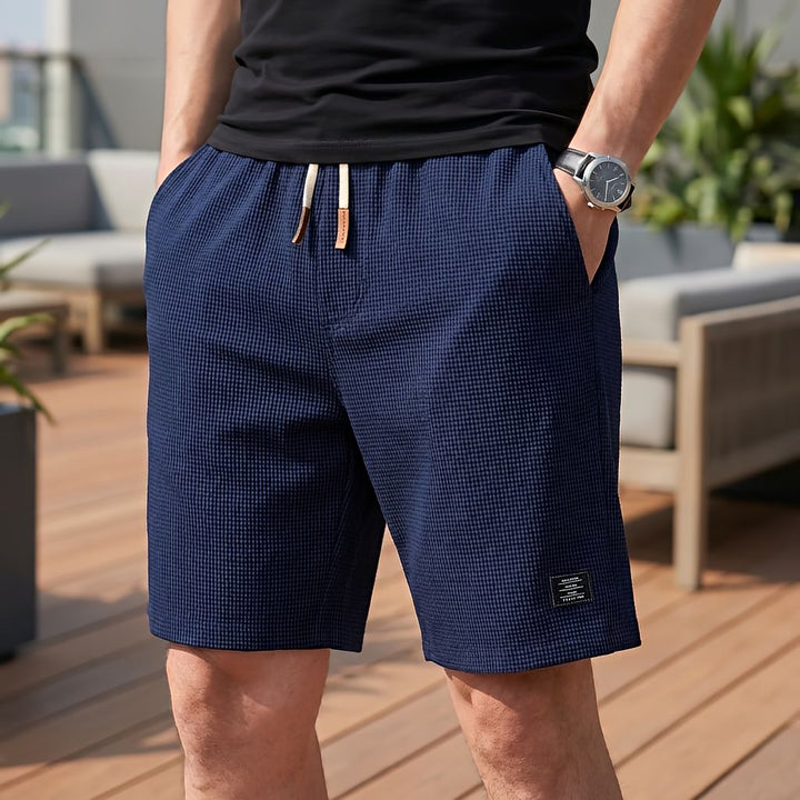 Mand i sort t-shirt og ur bærer mørkeblå vaffelstrikkede shorts med lyst snøre og lædertip, set forfra på en terrasse. 7