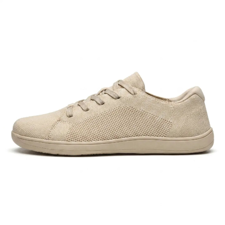 Beige sneaker i åndbart strikmesh med elastiske snørebånd og flad sål, set fra siden på hvid baggrund. 1