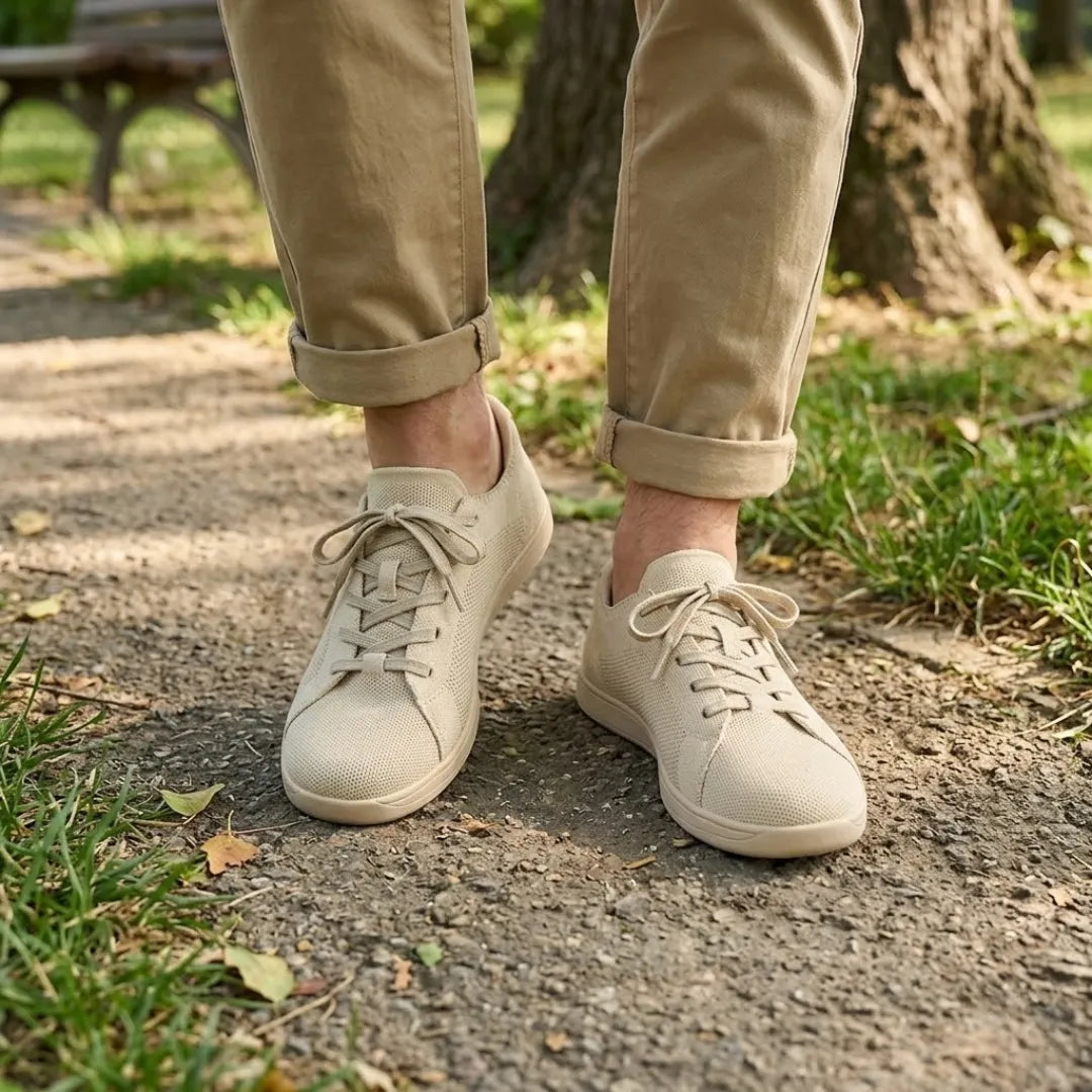 Beige strikmesh-sneakers båret af en person i oprullede khakibukser på en grussti i en park. 2