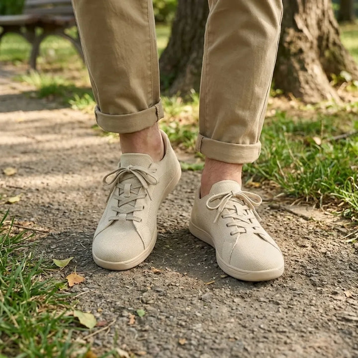 Beige strikmesh-sneakers båret af en person i oprullede khakibukser på en grussti i en park. 2