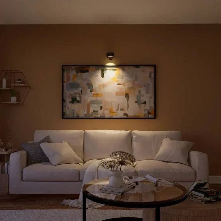 Sort væglampe oplyser et abstrakt maleri over en sofa i en stue om aftenen 15
