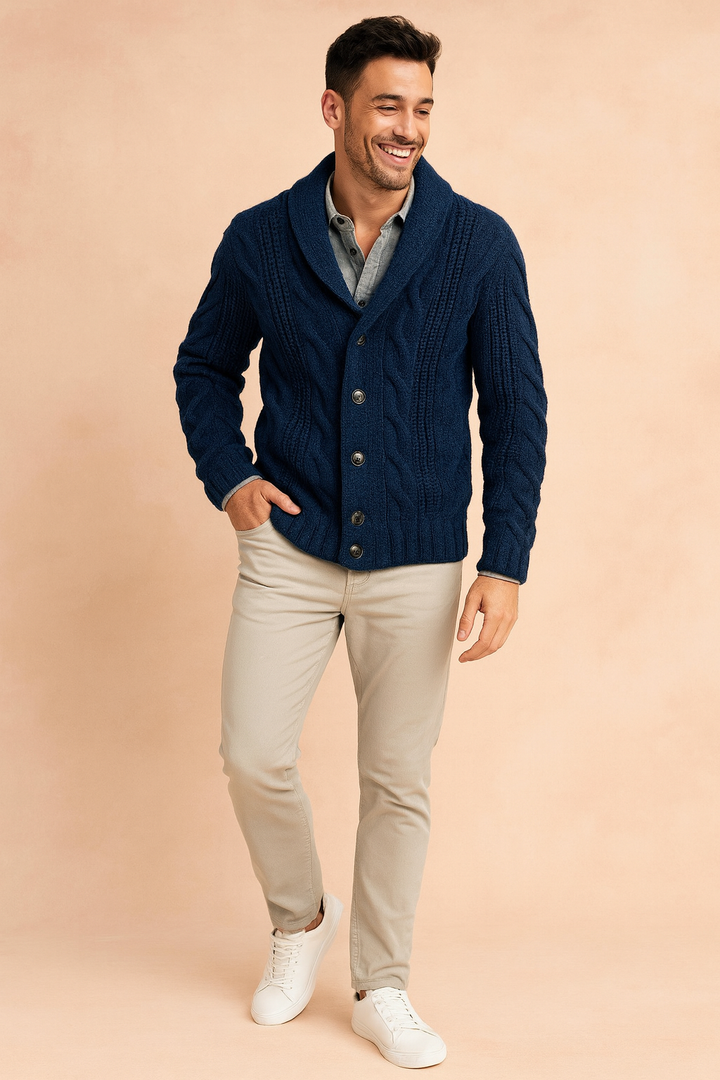 Helfigursbillede af mand i marineblå cardigan med flettemønster og sjalskrave, beige bukser og hvide sneakers, med hånden i lommen. 2
