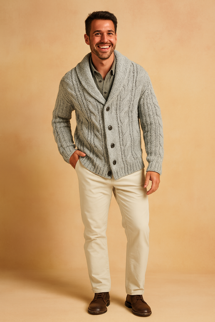 Helfigursbillede af mand i lysegrå flettestrikket cardigan med sjalskrave, cremefarvede bukser og brune snøresko, med en hånd i lommen. 8