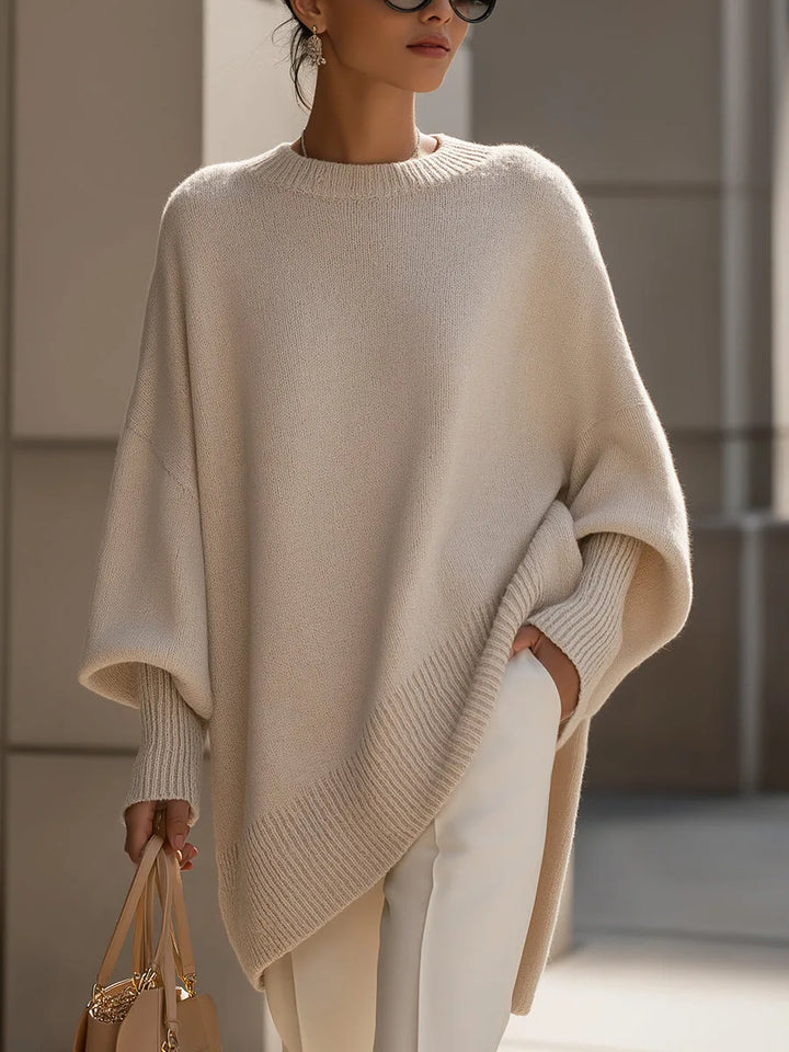 Model i beige oversize strikbluse med ribkant og hånden i lommen, hvide bukser og lille taske i hånden. 1