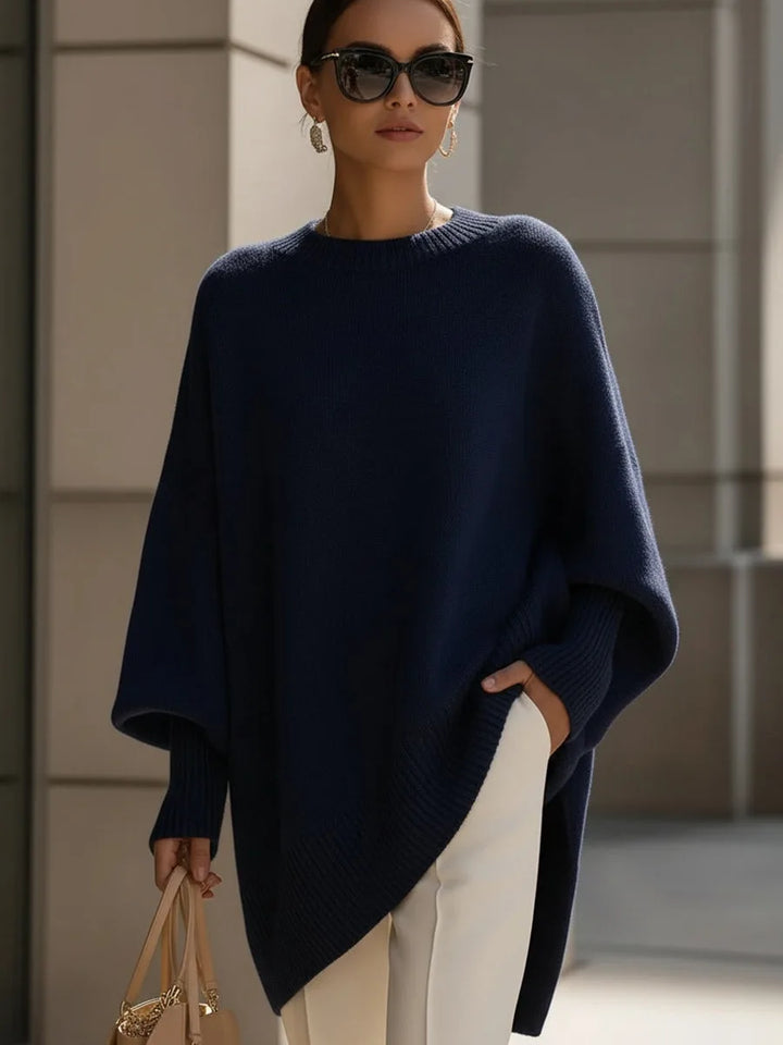 Model i mørkeblå oversize strikbluse med asymmetrisk kant, solbriller, hvide bukser og taske. 8