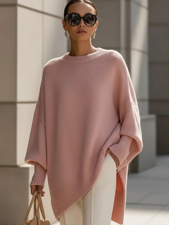 Model i lyserød oversize strikbluse med asymmetrisk ribkant, solbriller, hvide bukser og taske i hånden. 11