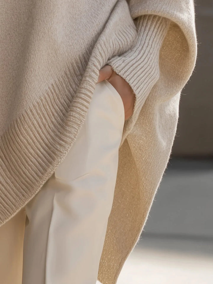 Nærbillede af sidesplit og den skrå ribkant på den beige strikbluse, med hånden i lommen på hvide bukser. 5