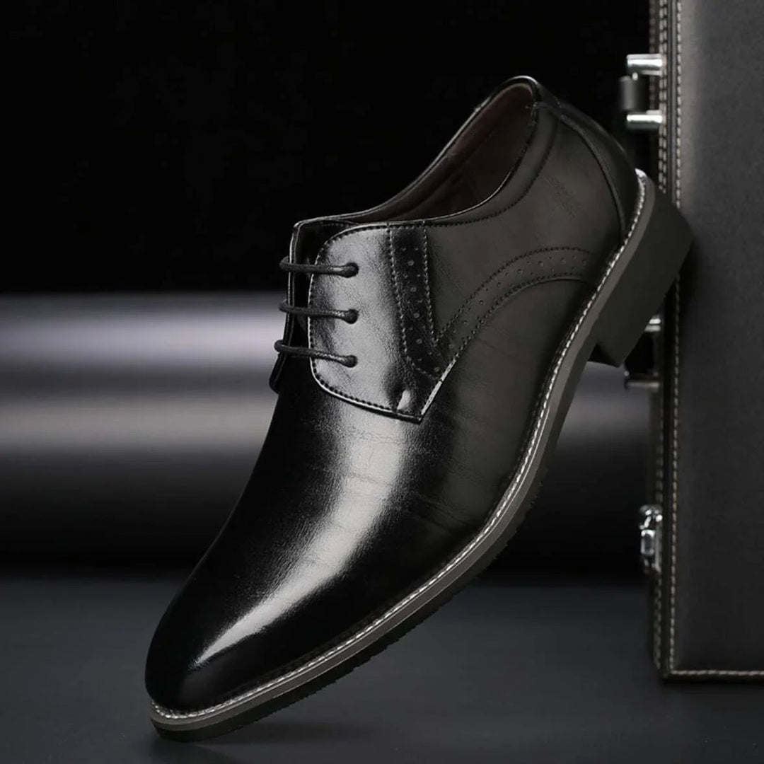 Sort, blank festsko med snørebånd og brogue-perforering, vist i sidebillede lænet mod en mørk kuffert. 3