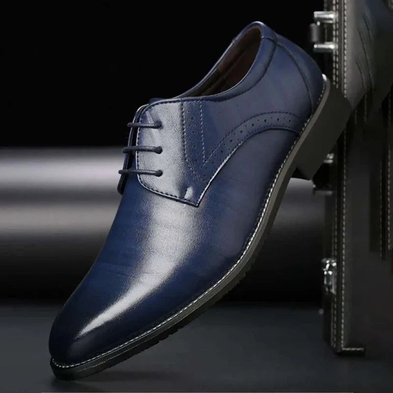 Mørkeblå, blank læderfestsko med snørebånd og brogue-perforering set fra siden, lænet mod en mørk kuffert. 5