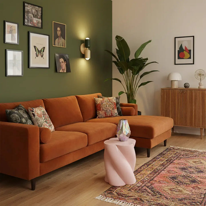Tændt væglampe i mørkegrøn og creme på grøn væg med indrammede billeder, over en rustorange hjørnesofa og et lyserødt sidebord. 9