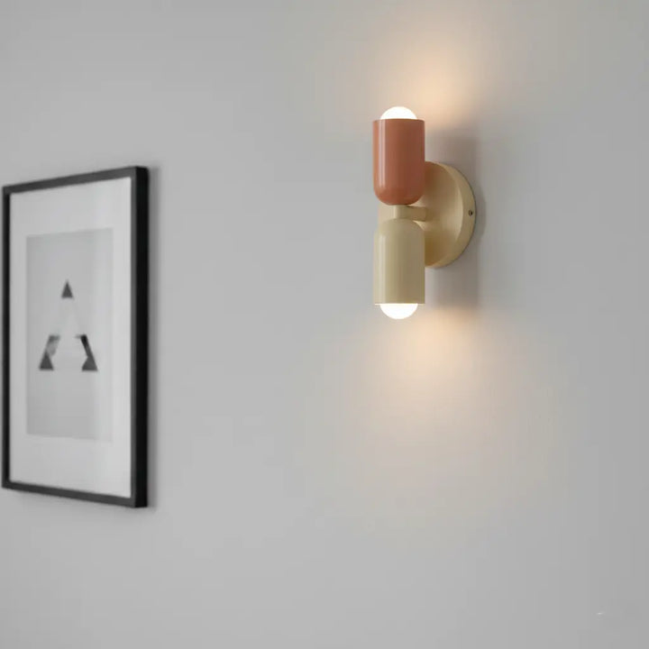 Tændt væglampe i terracotta og creme på lys grå væg, med lys op og ned og et indrammet grafisk billede ved siden af. 4