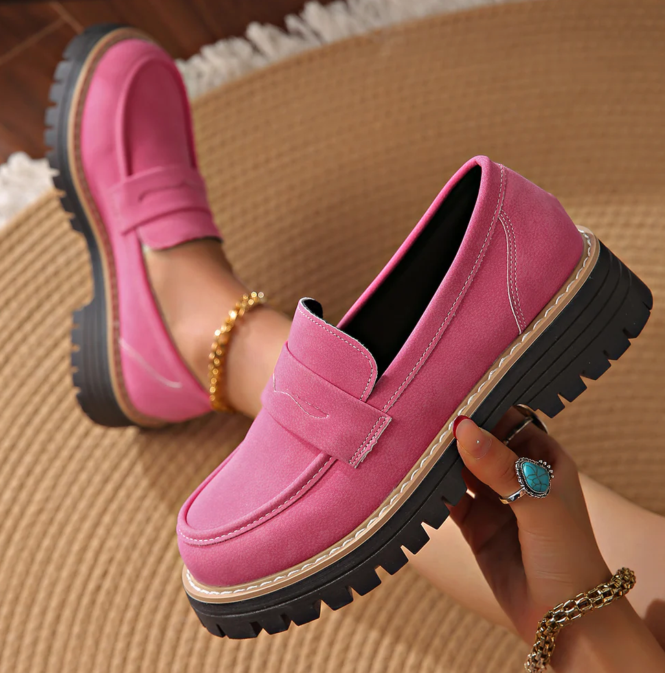 Pink ruskindslignende loafer holdt i hånden med turkis ring, mens den anden sko sidder på foden på et flettet tæppe. 18