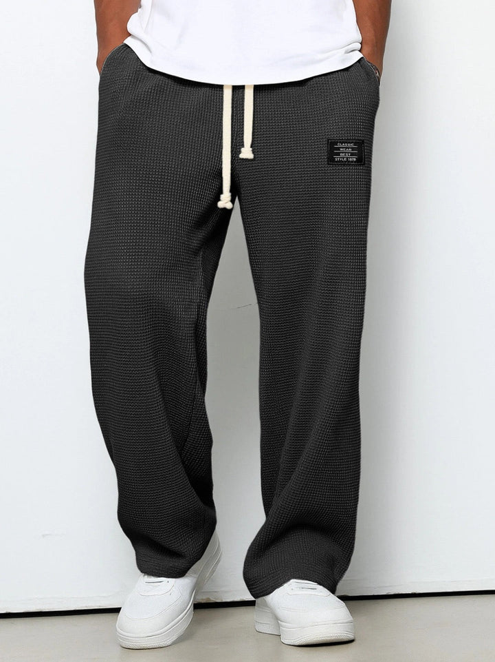 Mørkegrå vaffelstrikbukser med vide ben og hvidt snørebånd i taljen, båret af en model med hvid t-shirt og hvide sneakers. 1