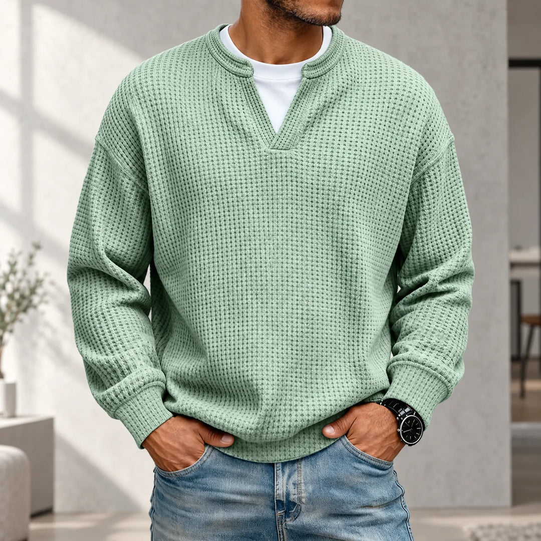Mand i lysegrøn vaffelstrikket trøje med lagdelt hvid T-shirt under kraven, hænderne i jeanslommerne, i lyst interiør. 1