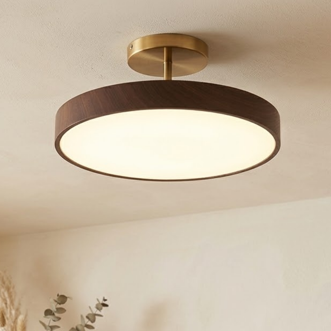 Tændt halvloftlampe med mørkt trælook og guldfarvet monteringsplade, set på skrå fra siden i et lyst rum med eukalyptusgren. 1