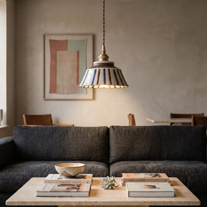 Tændt pendel med stribet keramisk skærm over et sofabord med bøger og skål, foran en mørk sofa i stuen. 5