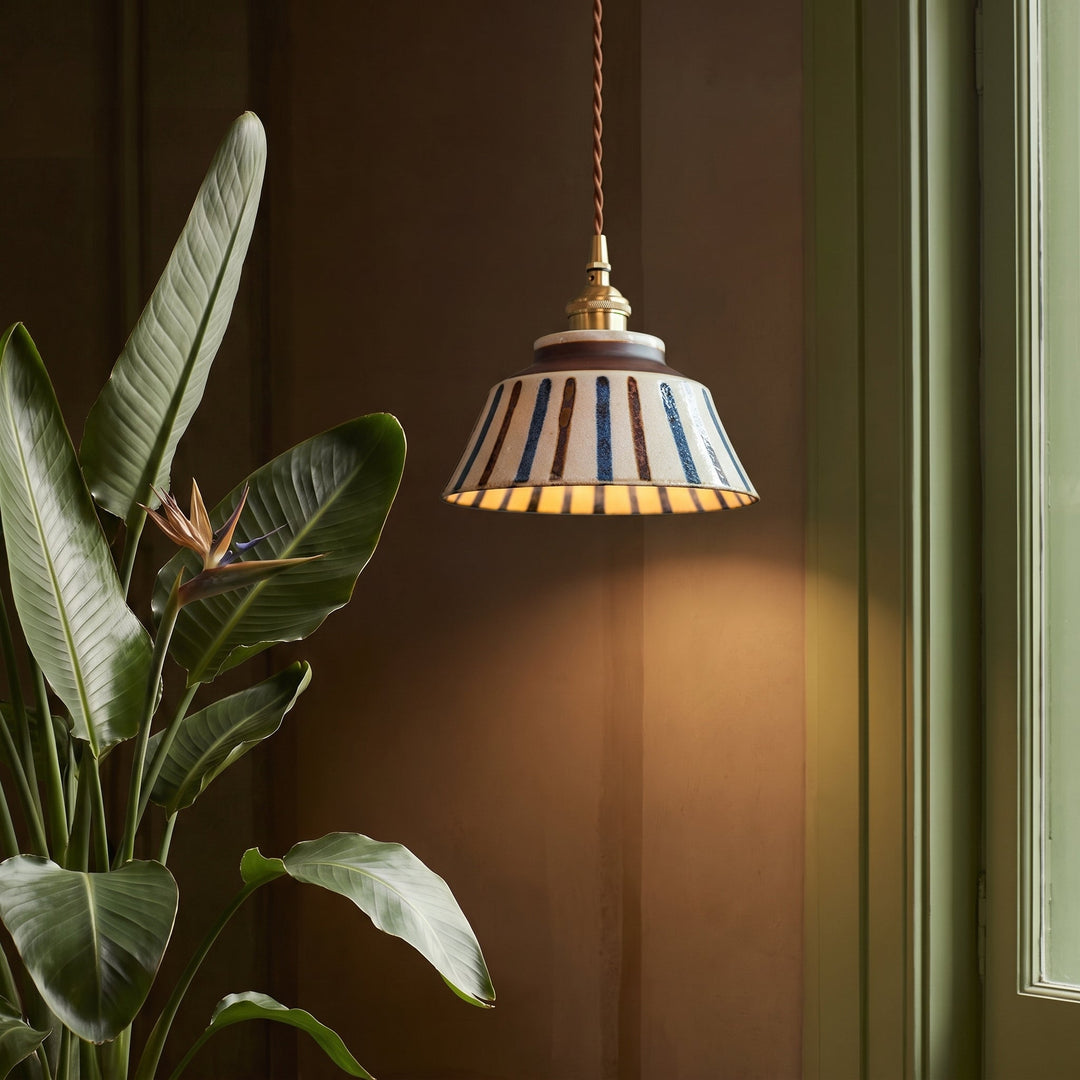Stribet keramisk pendel med messingfatning, tændt i et mørkt rum med stor grøn plante med blomst og grønt vindue. 4