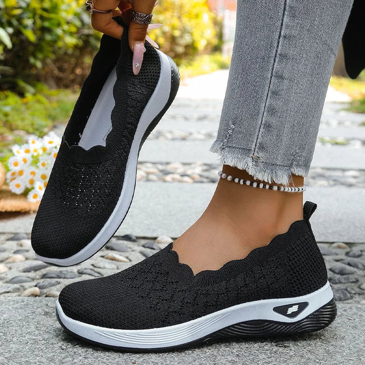 Sorte strikkede slip-on-sko med hvid mellemsål, den ene løftet i hånden og den anden på foden 13