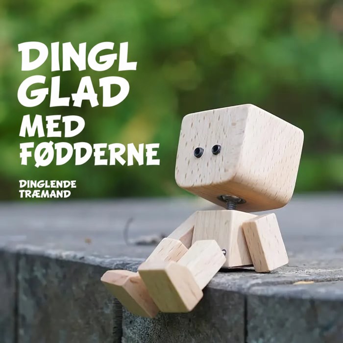 Træfigur med to små øjne sidder på en stenkant udendørs og lader benene dingle, foran grøn sløret baggrund. 15