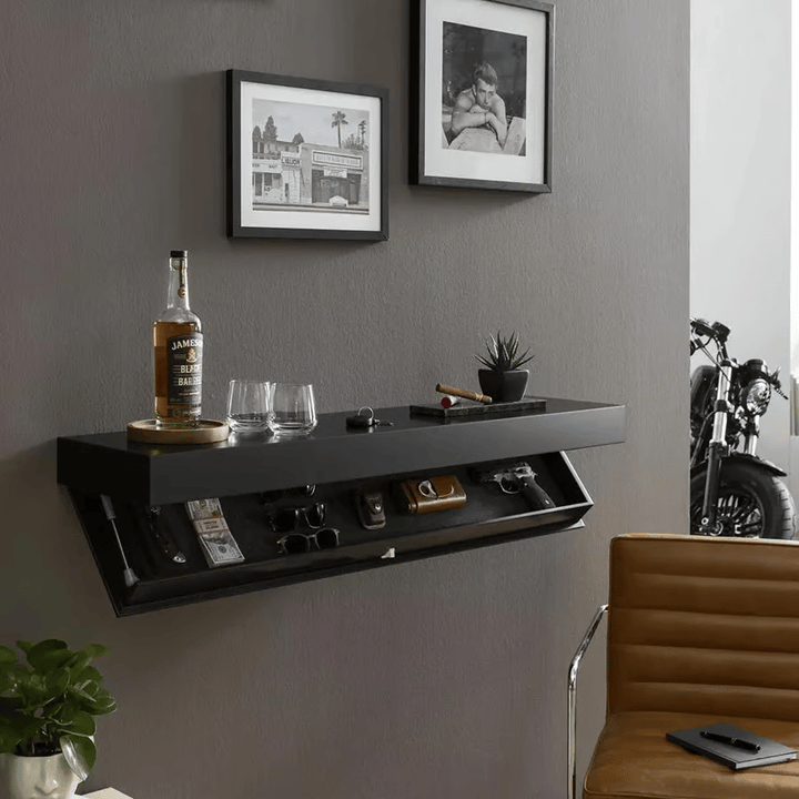 Sort svævende hylde på grå væg med åbent skjult rum og glas og flaske ovenpå, ved siden af indrammede fotos og en lædestol. 4