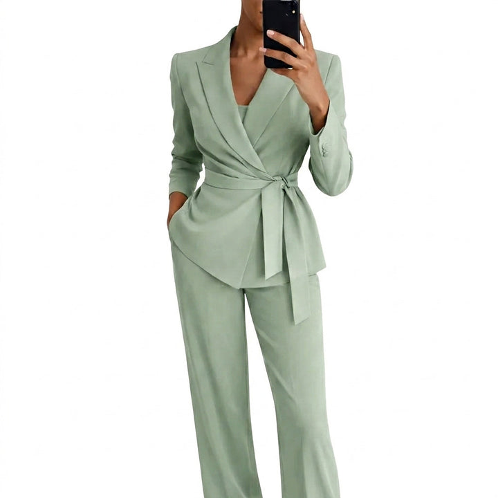 Model i lysegrønt sæt med blazer med omslagsfront og bindebælte samt vidde bukser, set forfra mens hun tager et spejlbillede med mobilen. 1