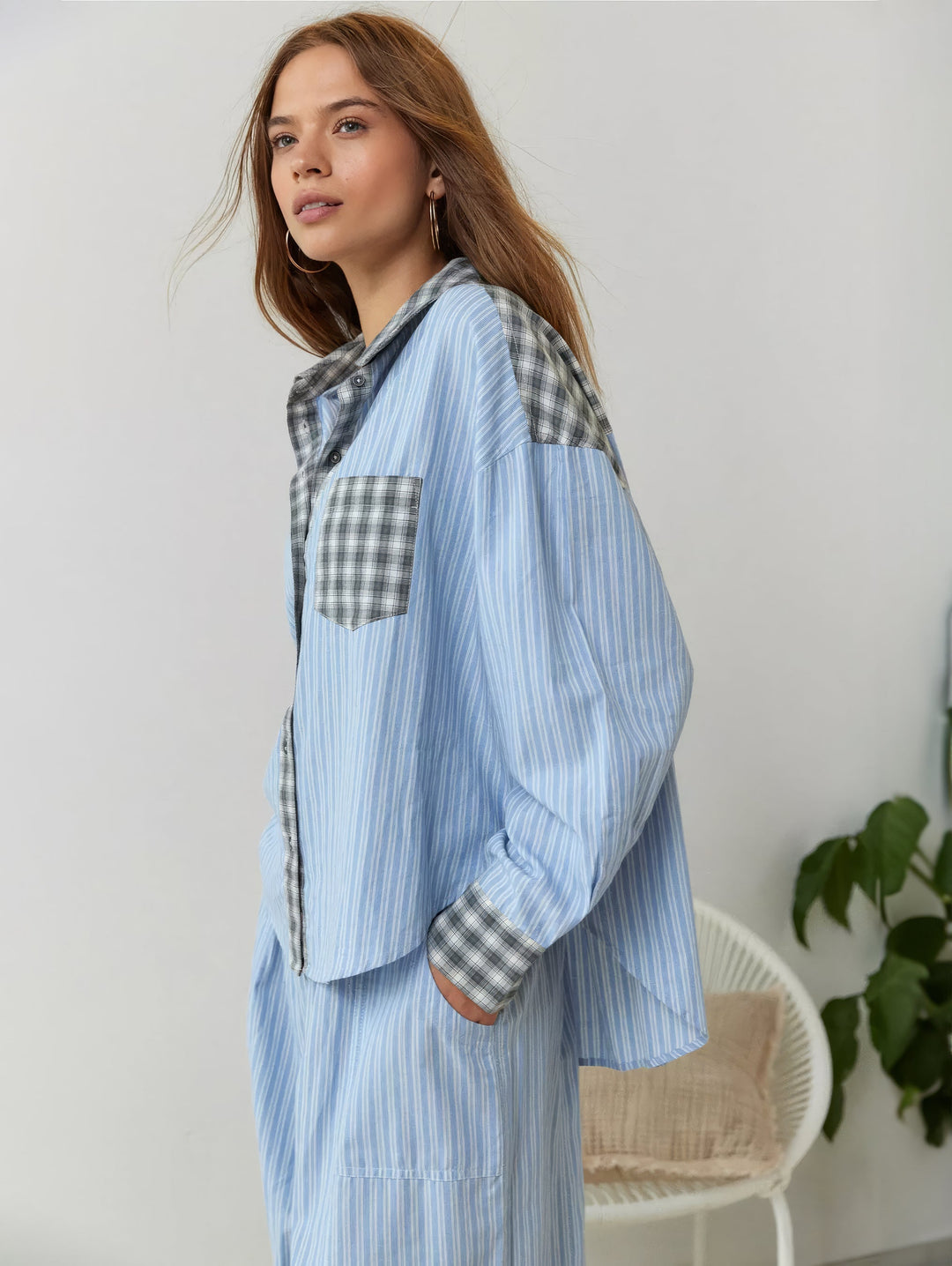 Model i blå stribet pyjamasskjorte med grå ternede detaljer, set fra siden foran en hvid kurvestol og en grøn plante. 3