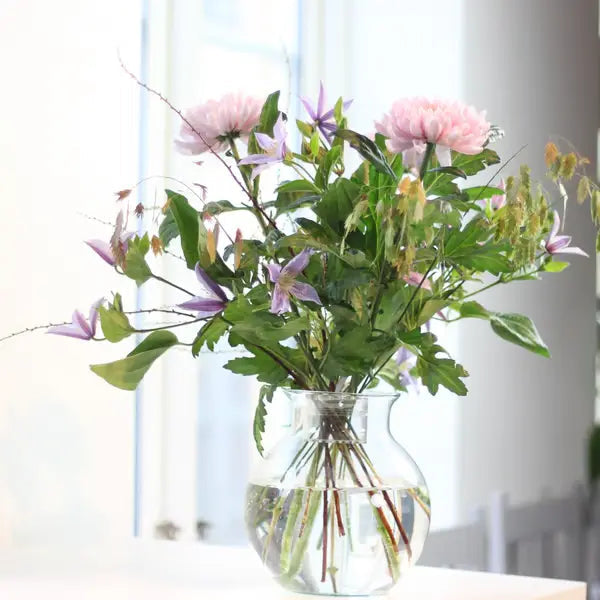 Rund glasvase med vand, hvor lyserøde dahlia, lilla blomster og grønne blade er samlet i en luftig buket ved et vindue. 4
