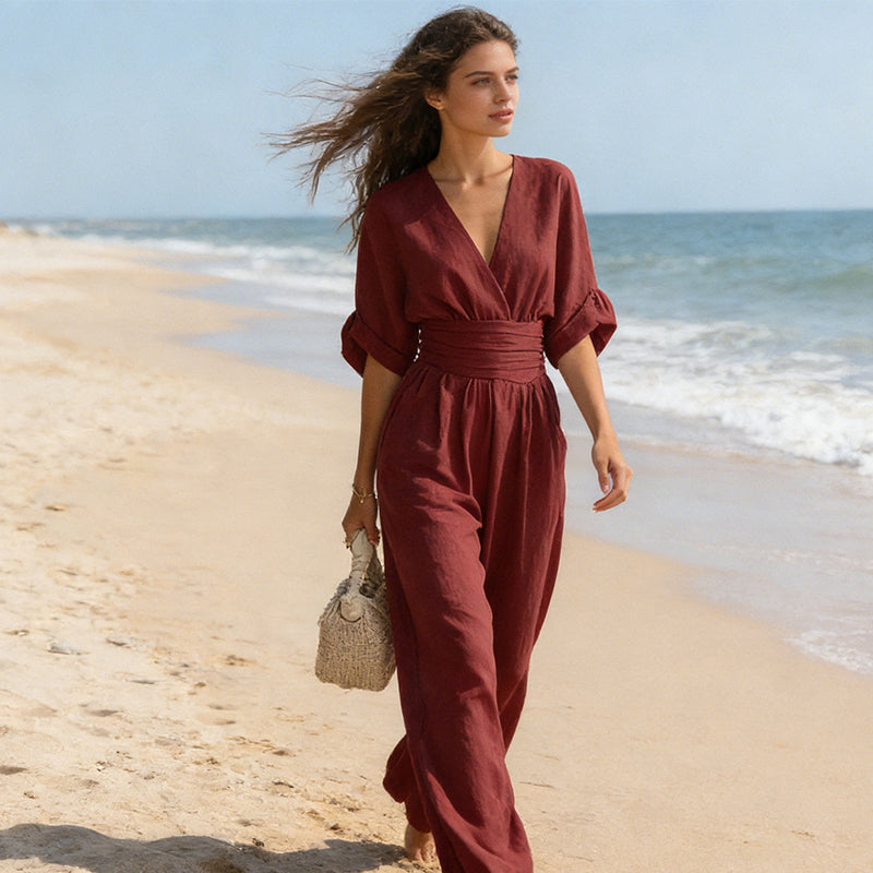 Model i mørkerød jumpsuit går barfodet langs stranden med håndtaske, mens vinden blæser i håret 7