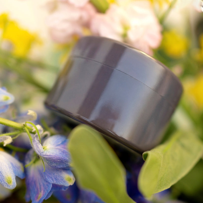 Mørkegrå, blank rund indsats set fra siden mellem blå blomster, grønne blade og sarte lyserøde og gule blomster. 10