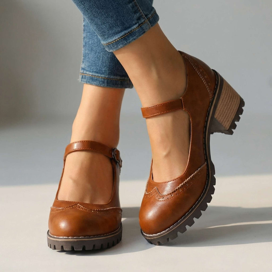 Brune Mary Jane sko med brogue-detaljer, kraftig sål og blokhæl, båret af model i jeans foran en lys baggrund. 2