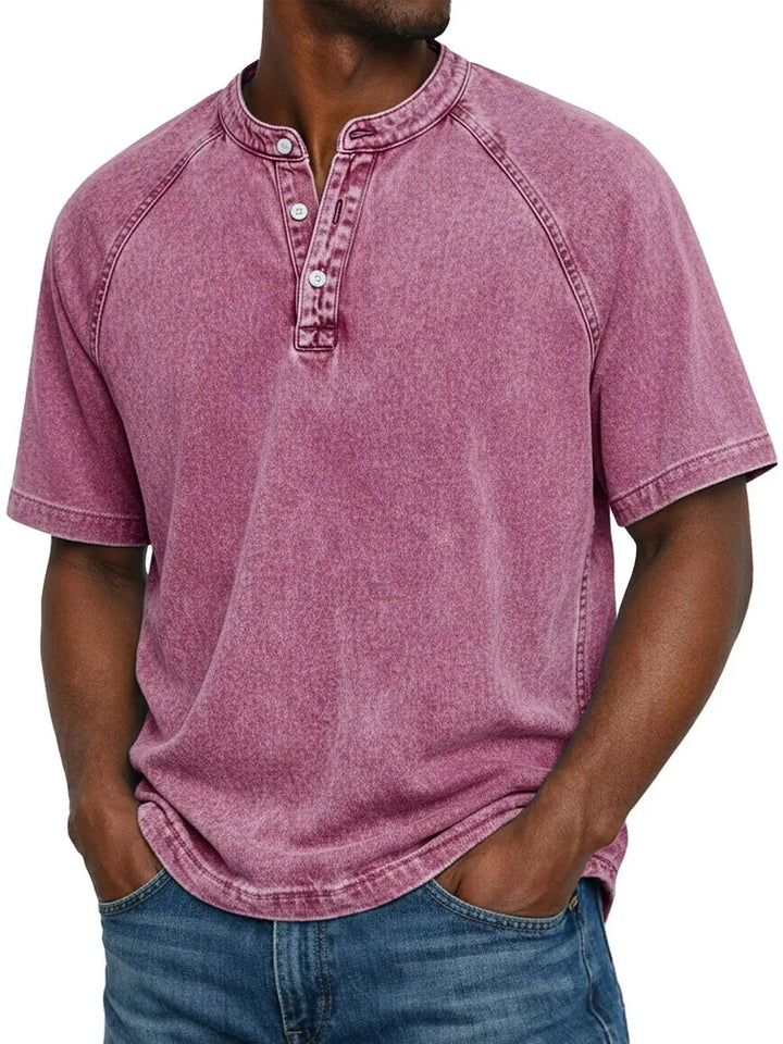 Mand i lyserød vasket Henley-T-shirt med raglanærmer og knaplukning ved halsen, hænderne i lommerne på jeans. 10