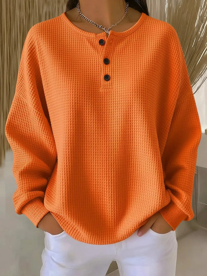 Ensfarvet orange vaffelstrikket trøje med tre sorte knapper ved halsen, båret med hvide bukser. 7