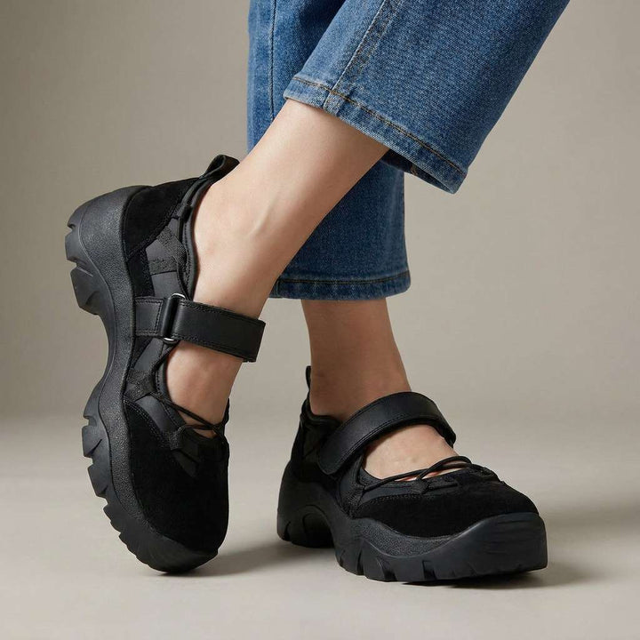 Helt sorte sko med velcrostrop og kraftig sål, båret med jeans på neutral studiebaggrund. 5