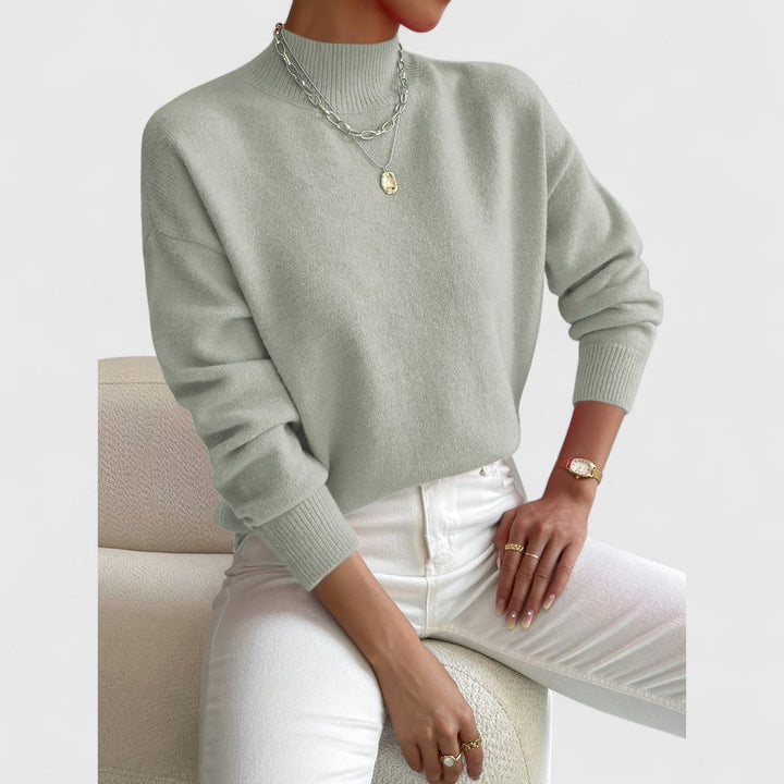 Model i lysegrøn strikbluse set lidt tættere på, siddende på en lænestol i hvide bukser med ringe og et armbåndsur. 3