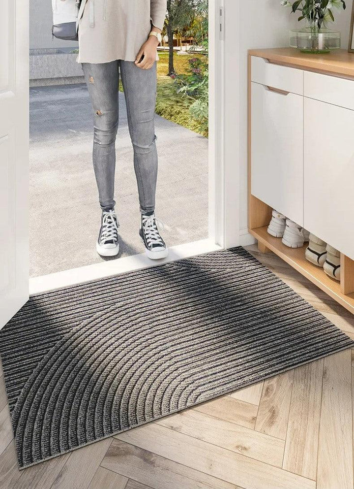Person i grå jeans og sneakers står udenfor døren, mens en stribet grå dørmåtte med buemønster ligger indenfor på fiskebensgulv. 5